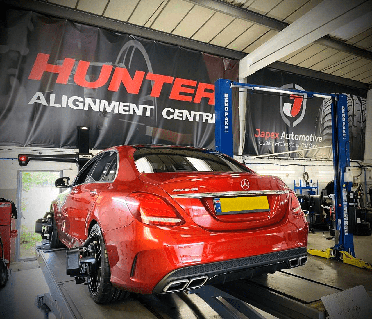 Image shows a red car facing away from us, on a wheel alignmenment ramp with a large black poster that says "Hunter Alignment Centre" on it.